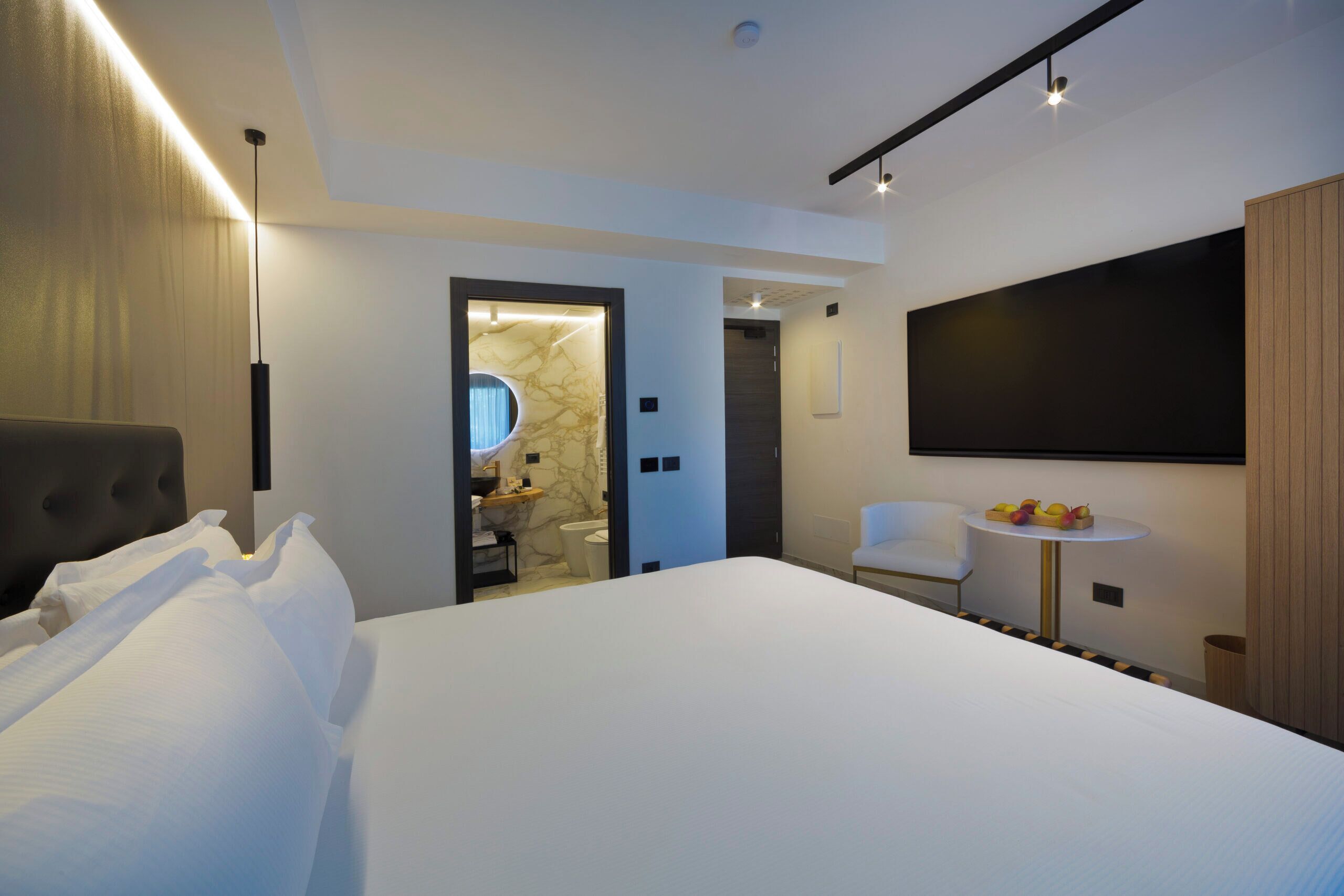 Et moderne hotelværelse med en stor seng, et fladskærms-tv på væggen, et lille rundt bord med frugt og et separat badeværelse, der kan ses gennem en åben dør.