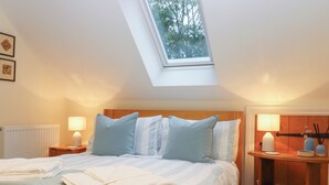 Ein Schlafzimmer mit Dachfenster, einem Bett mit gestreifter Bettwäsche, zwei Nachttischen mit Lampen und einem Holzbeistelltisch mit Büchern.