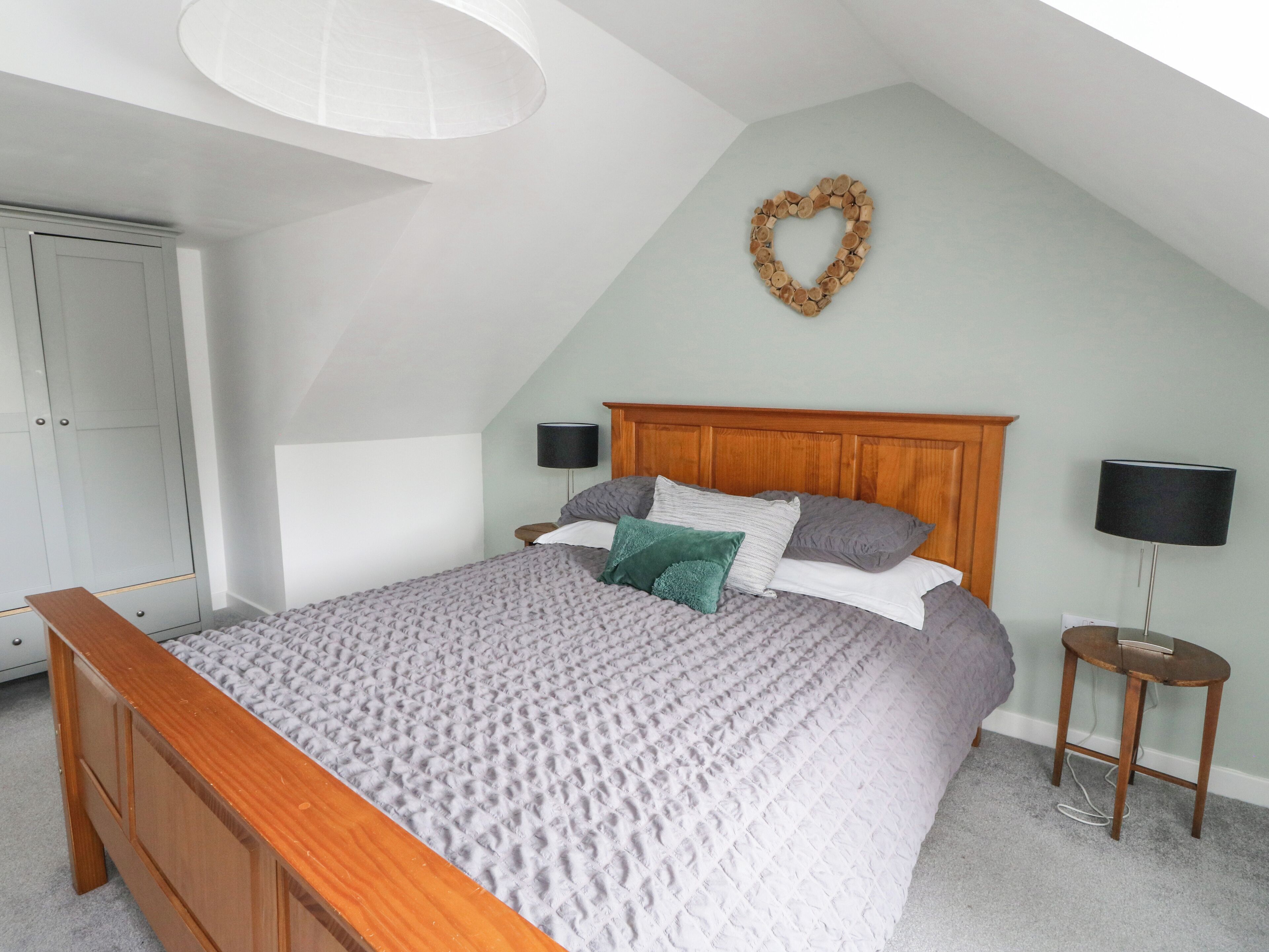 Une chambre avec un plafond en pente, un cadre de lit en bois, une couvre-lit matelassée grise, deux tables de chevet avec des lampes, une armoire et une décoration en forme de cœur accrochée au mur.