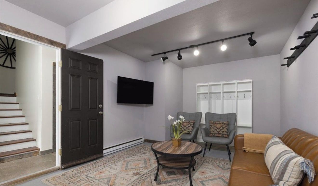 A living room with a sofa, two armchairs, a coffee table, a flat-screen TV, a door, and a staircase.