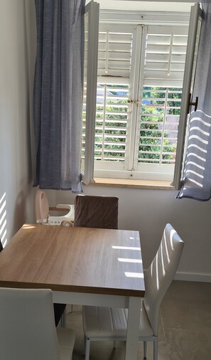 Una habitación con una mesa de madera, sillas blancas, una ventana con contraventanas blancas y vistas al exterior con vegetación.