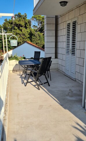 Un patio con una mesa y sillas, una cesta colgante, y un edificio con una puerta y contraventanas blancas.