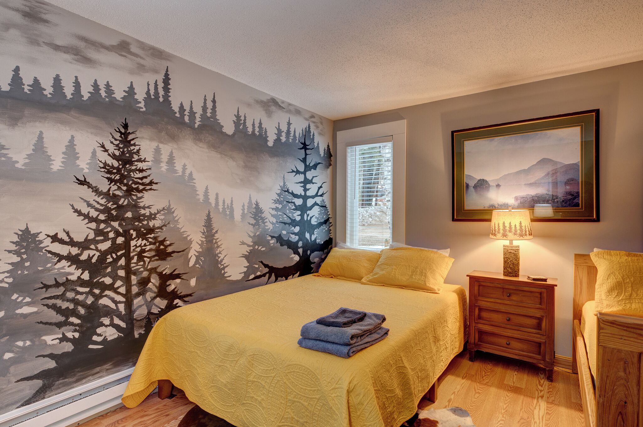 A bedroom with a forest mural, a bed with yellow bedding, a nightstand with a lamp, and a window with blinds.