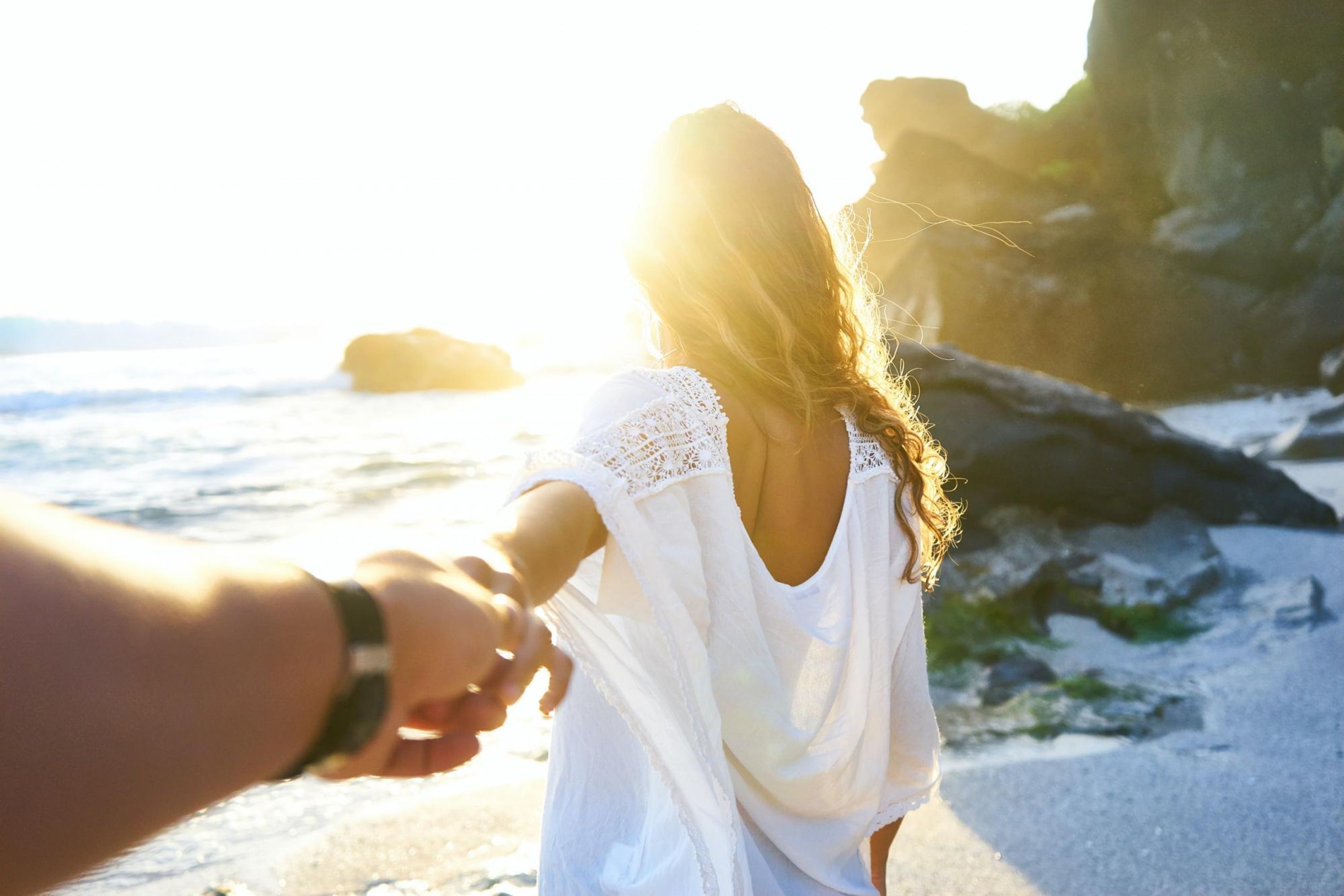 A couple holding hands on a beach with a rocky shoreline and ocean in the background.