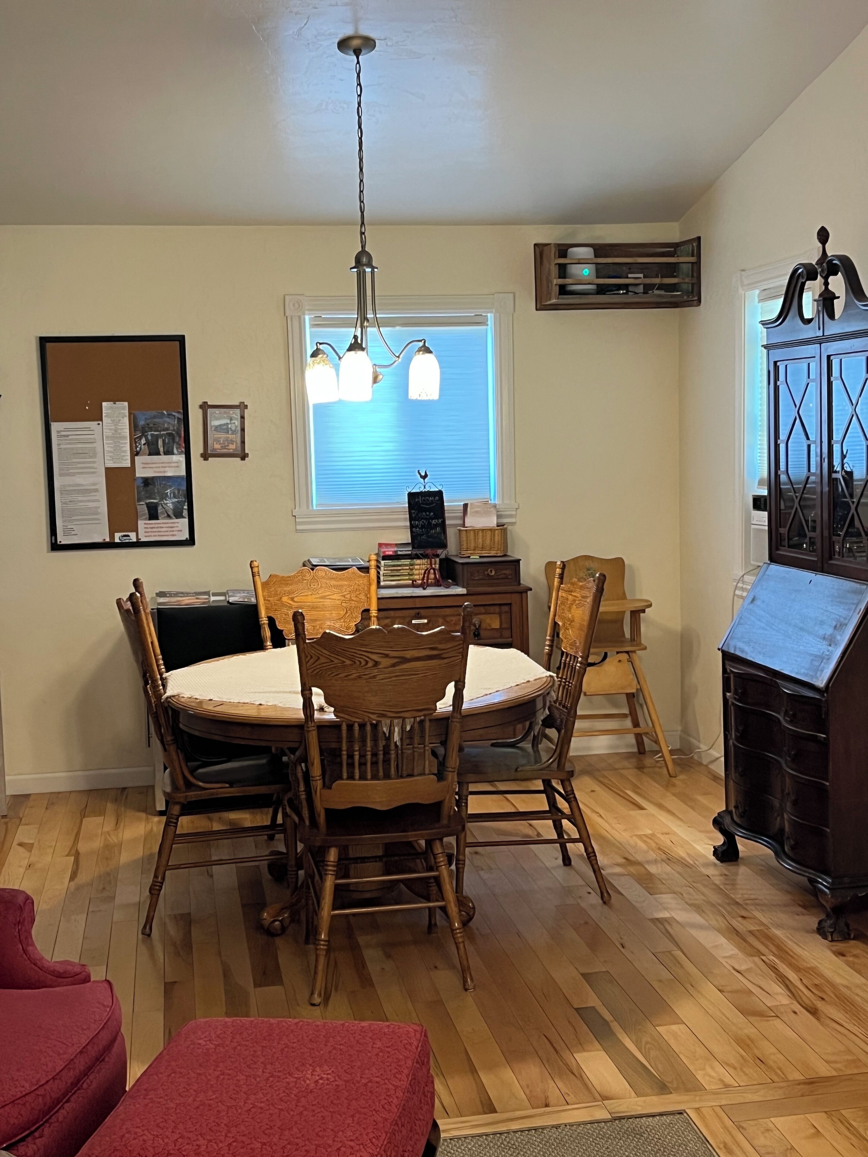 A dining area with a wooden table and chairs, a hanging light fixture, a bulletin board, and a window with blinds.