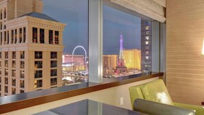 Ein Hotelzimmer mit großem Fenster, das einen Blick auf eine nächtliche Stadtlandschaft bietet, darunter ein markanter Turm und ein Riesenrad.