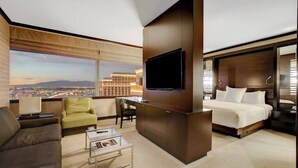 A modern hotel room with a large TV, a sofa, a chair, a small table, and a bed.