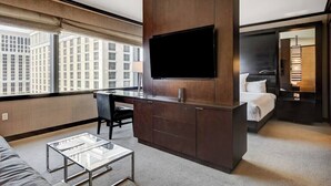 Une chambre d’hôtel moderne dotée d’un grand téléviseur fixé sur un meuble en bois, d’un bureau avec une chaise, d’un lit avec une table de chevet et offrant une vue sur les bâtiments de la ville à travers de grandes fenêtres.