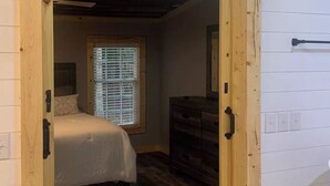 Une chambre avec une porte en bois, un lit, une commode et une fenêtre avec des stores.