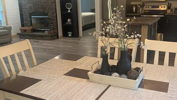 Une salle à manger avec une table dressée pour quatre personnes, une cheminée, une cuisine équipée d’appareils modernes et un salon avec un canapé et un téléviseur.