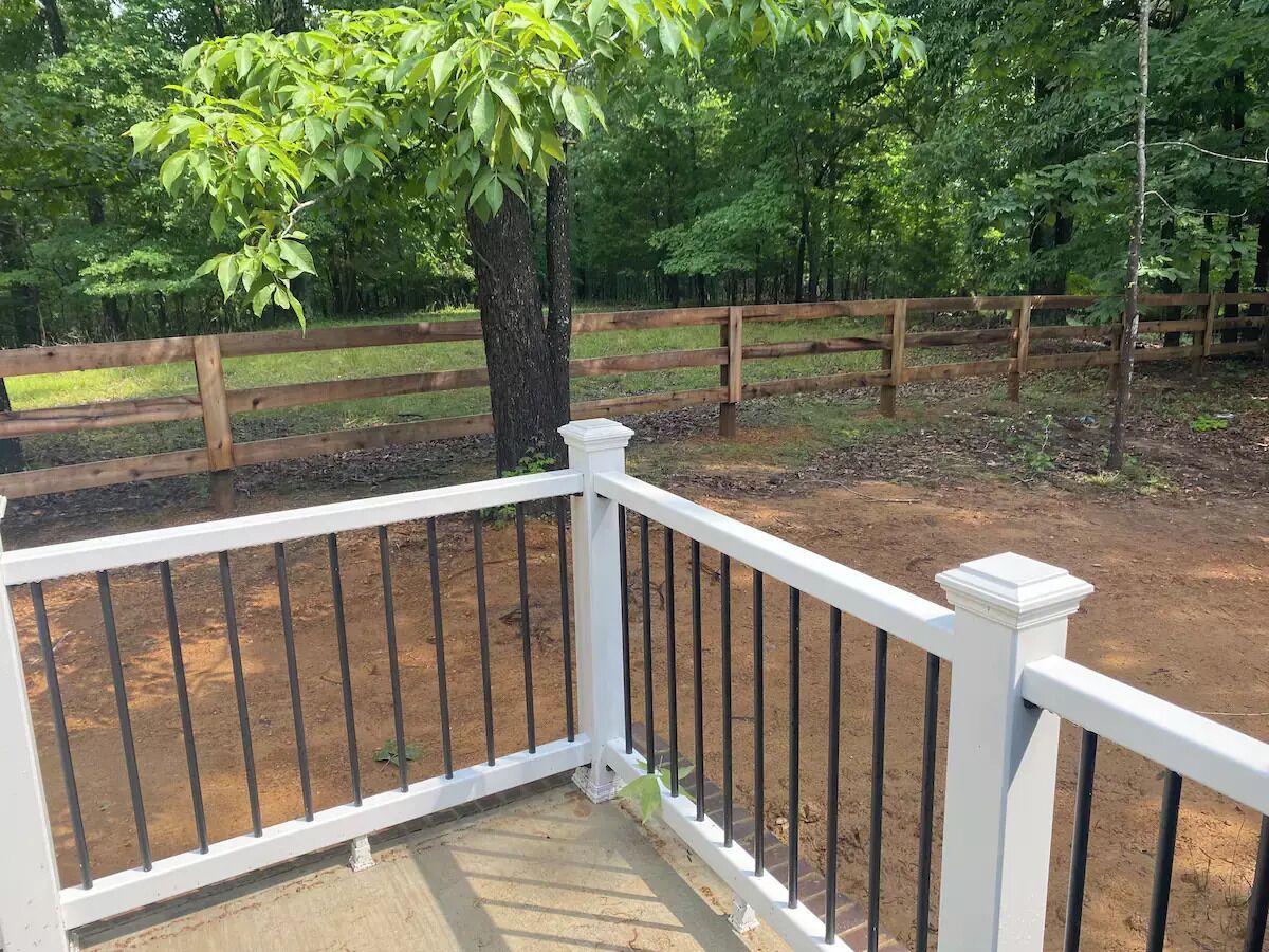 A wooden deck with a white railing, surrounded by a dirt area and a wooden fence.
