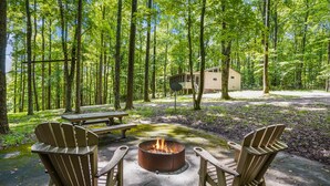 A forested campsite with a fire pit, picnic table, and Adirondack chairs.