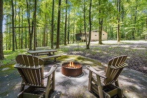 Een bebost kampeerterrein met een vuurkuil, picknicktafel en Adirondack stoelen.