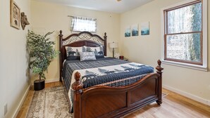A bedroom with a wooden bed, a ceiling fan, a window with a forest view, and a wall-mounted clock. - Woodland Cottage | Secluded Hocking Hills Getaway (Laurelville)