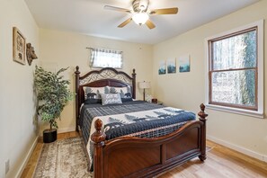 Une chambre avec un lit en bois, un ventilateur de plafond, une fenêtre donnant sur la forêt, une table de chevet avec une lampe et une plante.