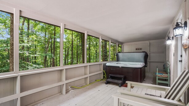 A room with a hot tub, wooden deck, and large windows overlooking a forest.