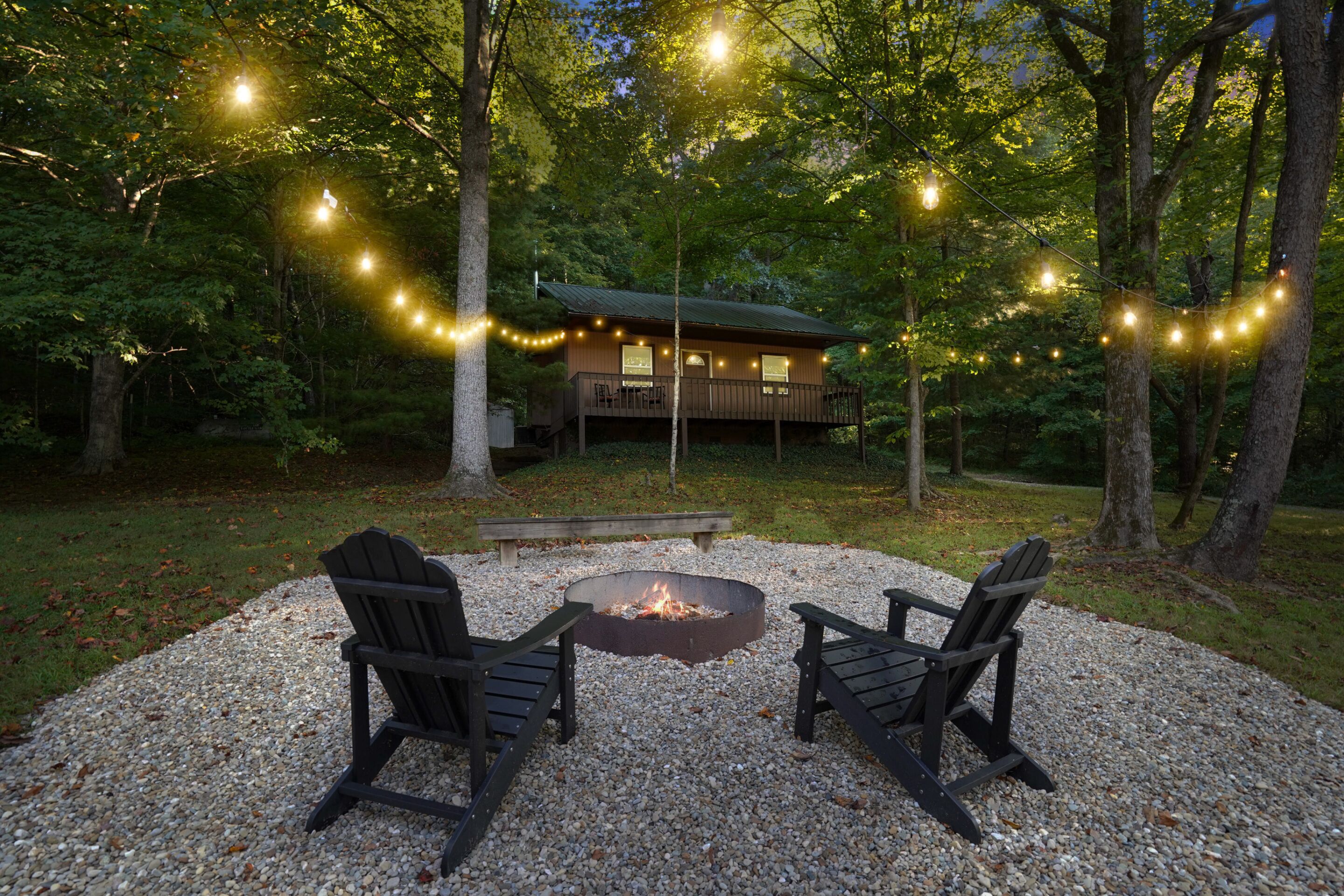 A cabin with a deck, surrounded by trees, and a fire pit with two chairs in front.