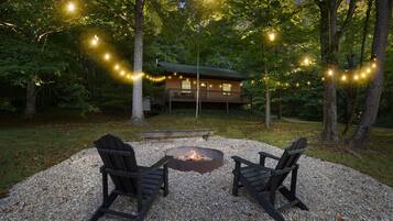 A cabin with a deck, surrounded by trees, and a fire pit with two chairs in front.