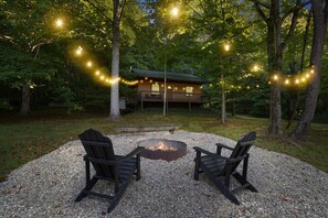A cabin with a deck, surrounded by trees, and a fire pit with two chairs in front.