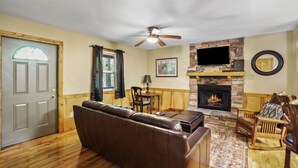 A living room with a fireplace, leather sofa, wooden flooring, and a television mounted above the fireplace. - Outback Cabin | Hocking Hills, Cozy Cabin for 2 (Laurelville)