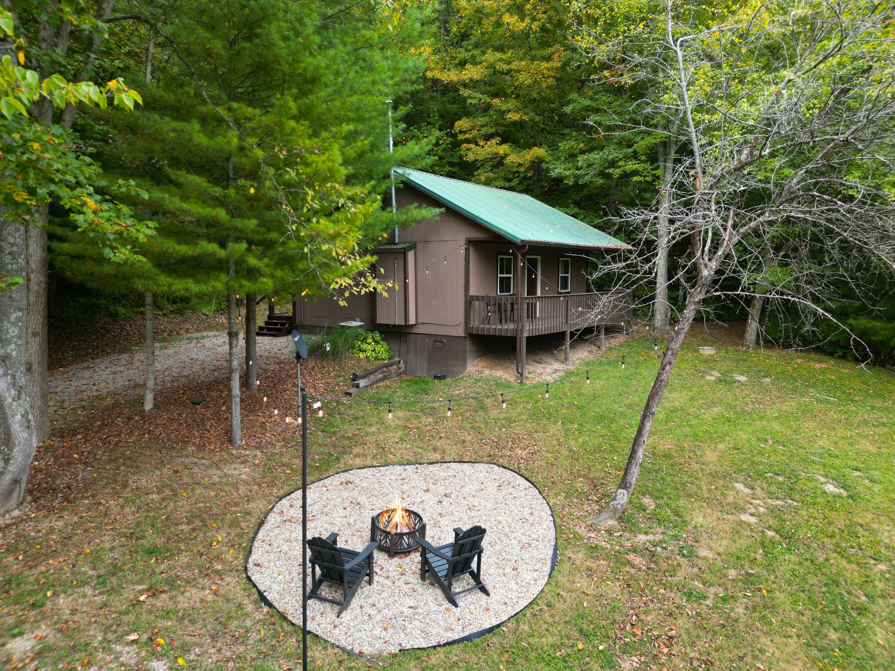 A cabin with a deck, surrounded by trees and a fire pit area with chairs.