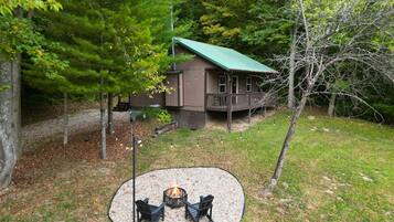 A cabin with a deck, surrounded by trees and a fire pit area with chairs.