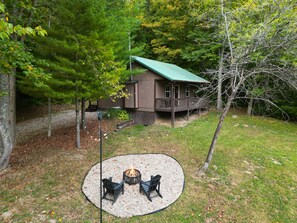 A cabin with a deck, surrounded by trees and a fire pit area with chairs. - The Hideout in Hocking Hills | Cozy Couples Escape (Laurelville)