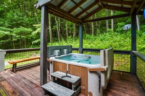 A wooden deck with a hot tub, benches, and a covered area surrounded by forest. - The Hideout in Hocking Hills | Cozy Couples Escape (Laurelville)