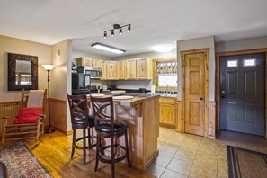 A kitchen with wooden cabinets, a central island, bar stools, a microwave, and a refrigerator. - The Hideout in Hocking Hills | Cozy Couples Escape (Laurelville)
