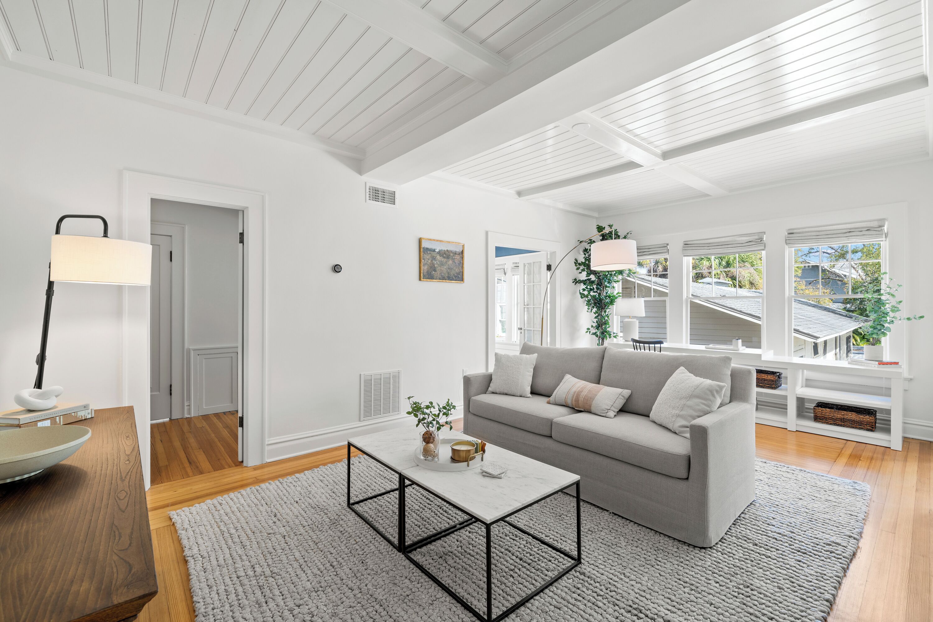 A modern living room with a white sofa, a coffee table, and a lamp. There are windows with blinds, a door, and a plant on the windowsill.