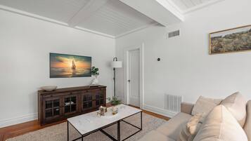 A living room with a white ceiling, a wooden cabinet, a white coffee table, a beige sofa, and a painting of a sunset over the ocean.