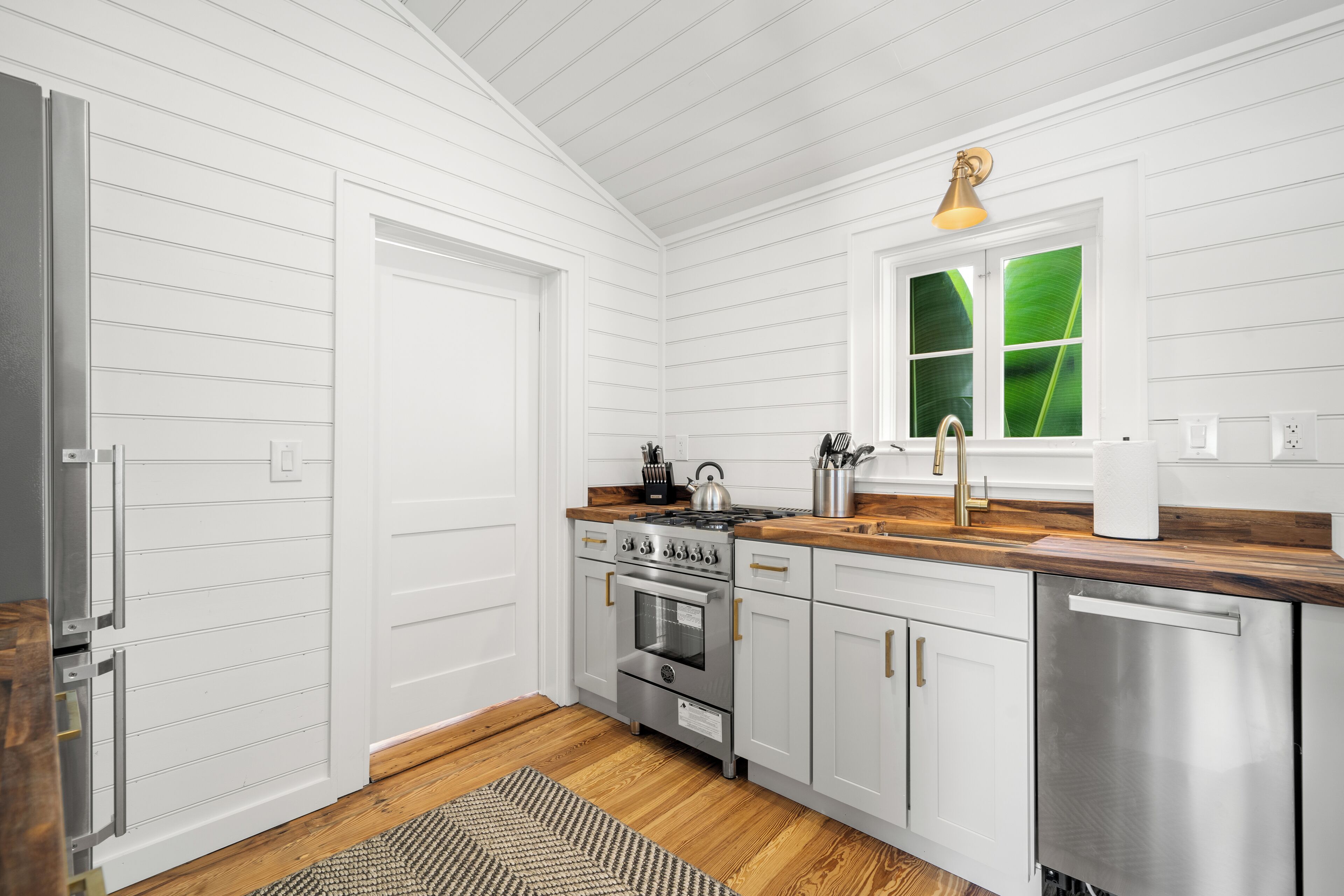 A kitchen with white paneling, a wooden countertop, stainless steel appliances, and a window with a view of greenery.