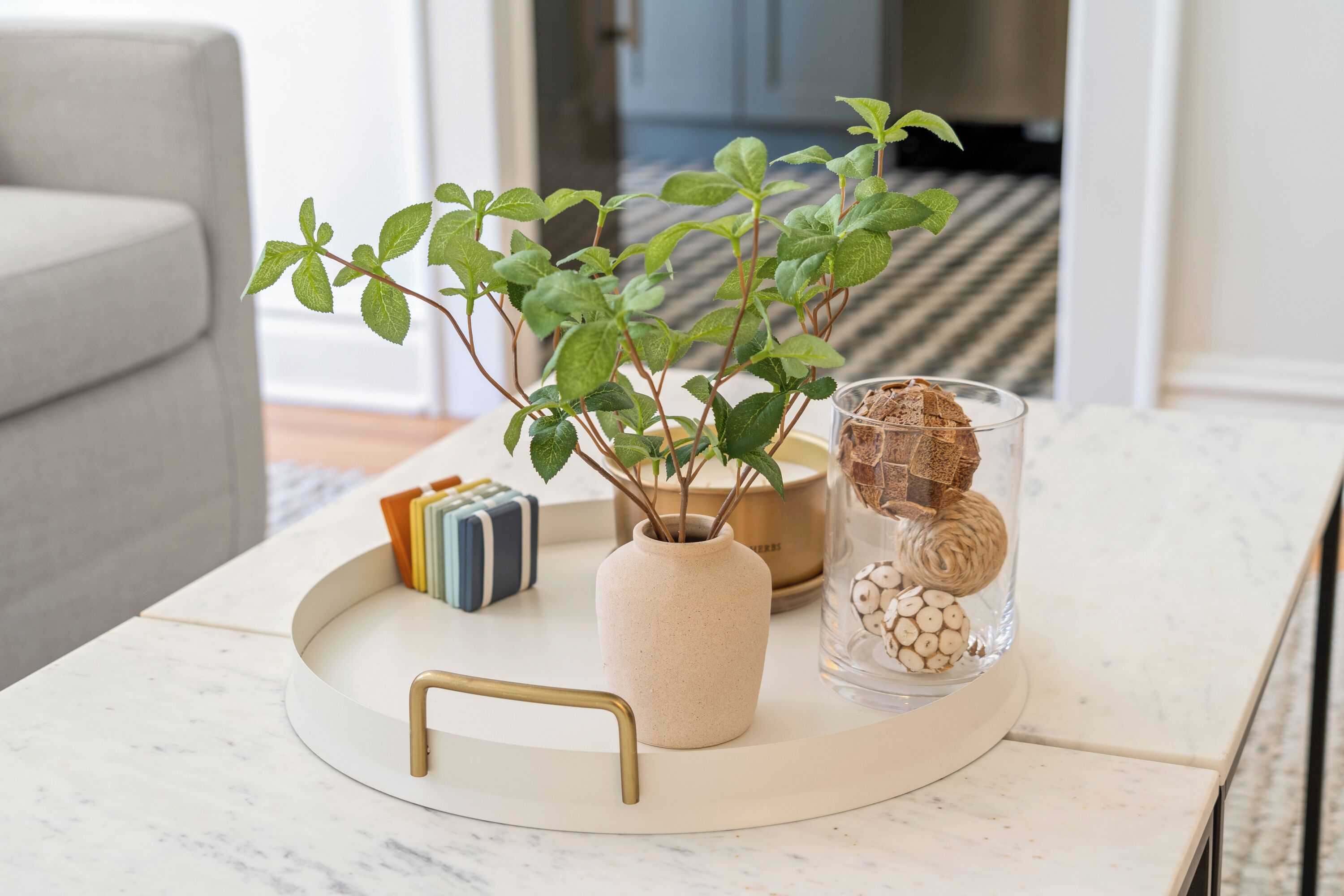 A round tray on a marble table with a beige vase, a glass jar with decorative items, and a small plant.