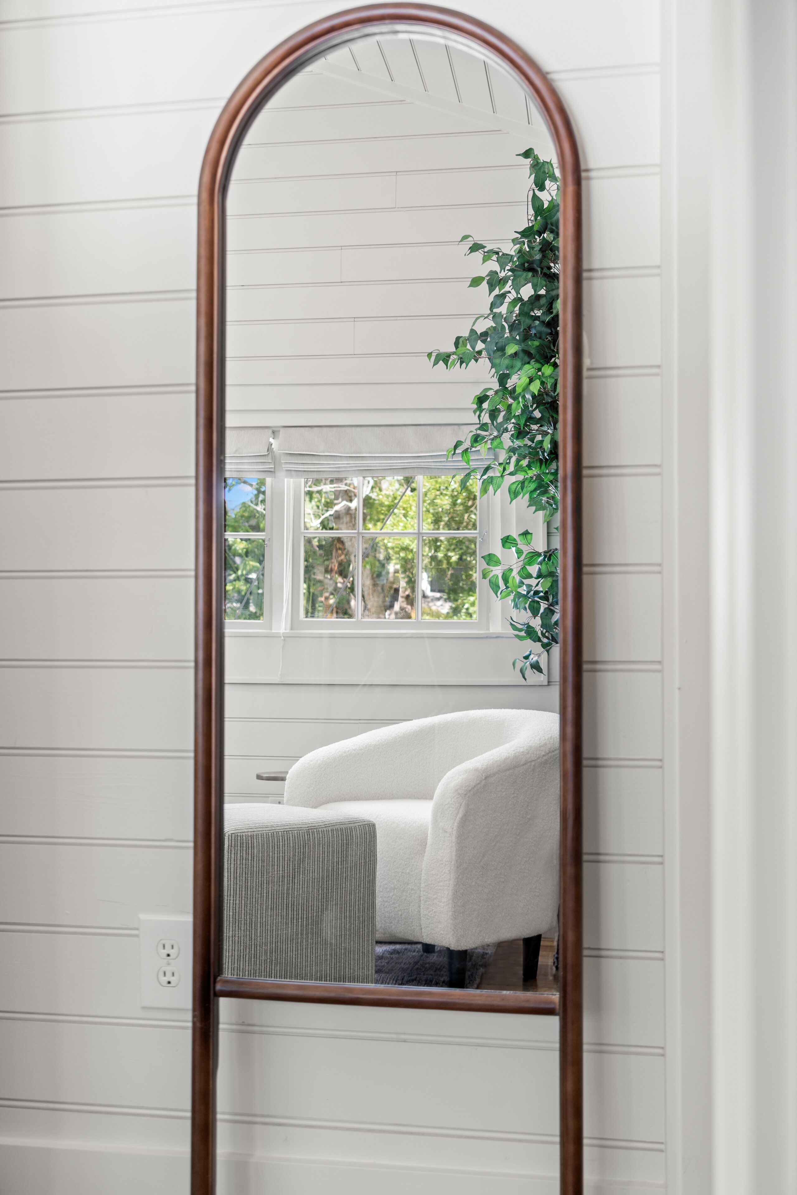 Ein Spiegel mit Holzrahmen, der einen Raum mit weißen Wänden, einem Fenster und einer Pflanze reflektiert.