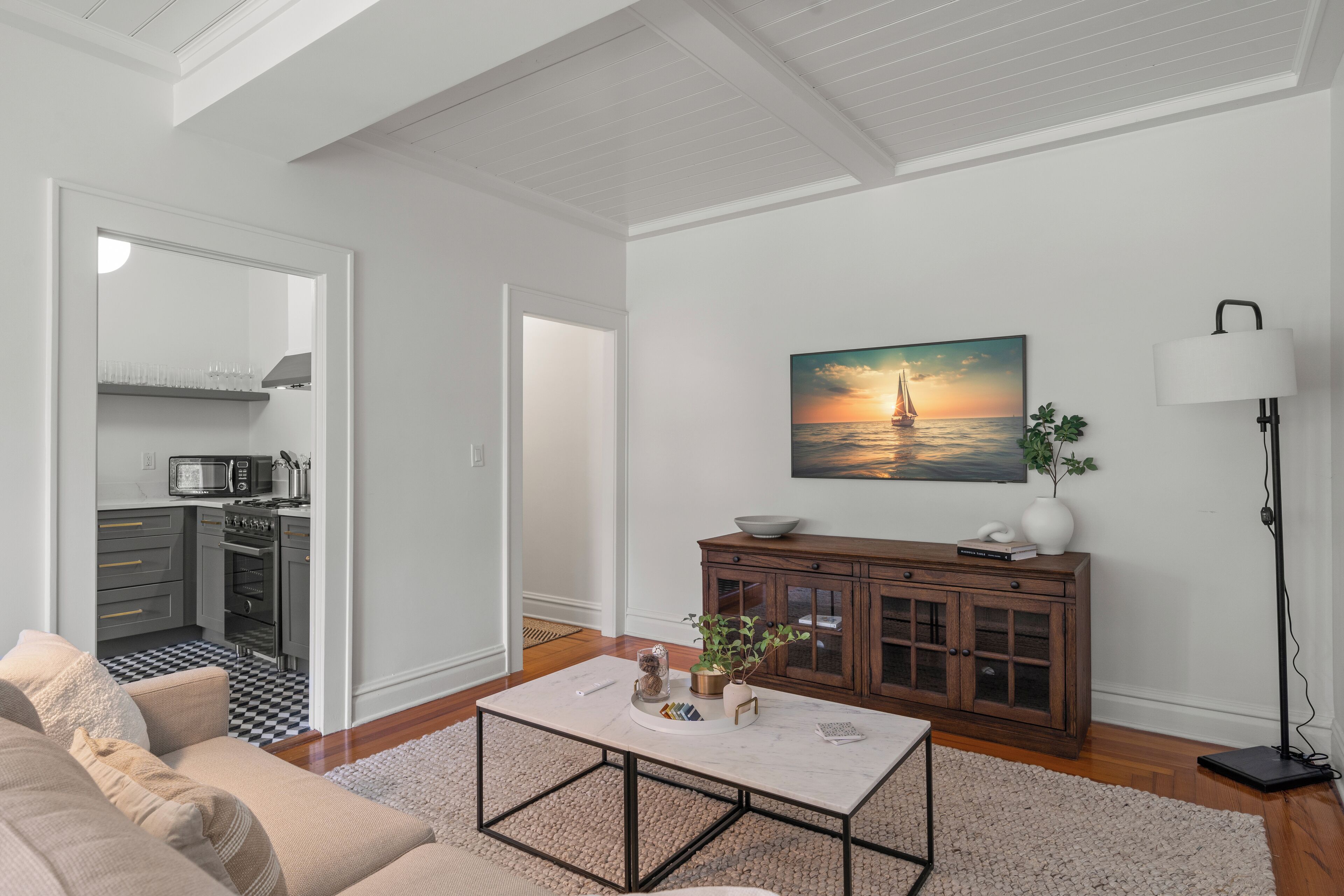 A living room with a sofa, coffee table, and a painting of a sunset over water. There is a cabinet with decorative items and a floor lamp.