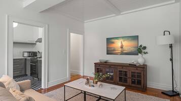 A living room with a sofa, coffee table, and a painting of a sunset over water. There is a cabinet with decorative items and a floor lamp.