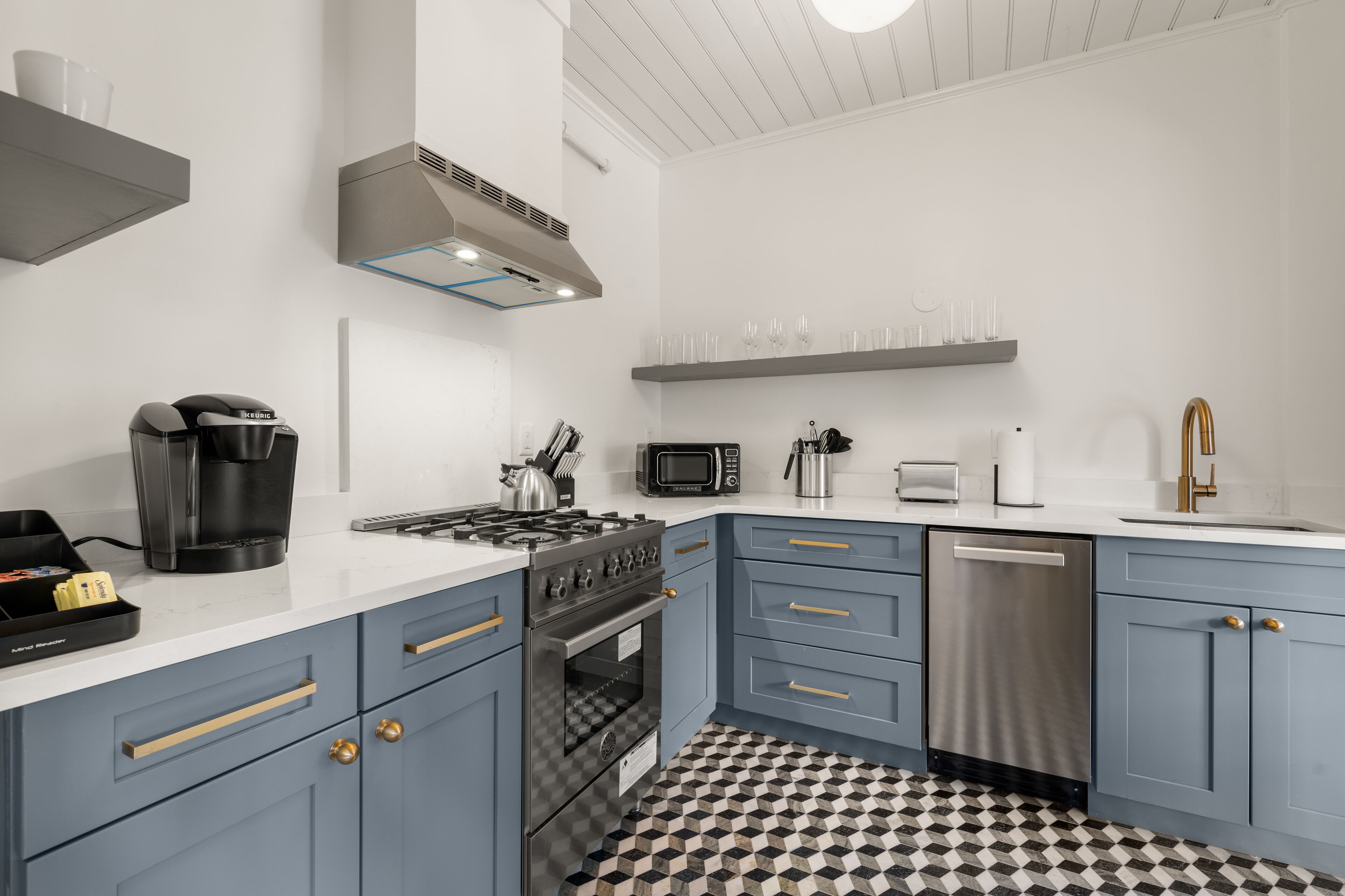 Una cocina moderna con estufa a gas, horno, microondas y lavavajillas. Hay armarios azules con detalles dorados, una encimera blanca y un suelo a cuadros blancos y negros.