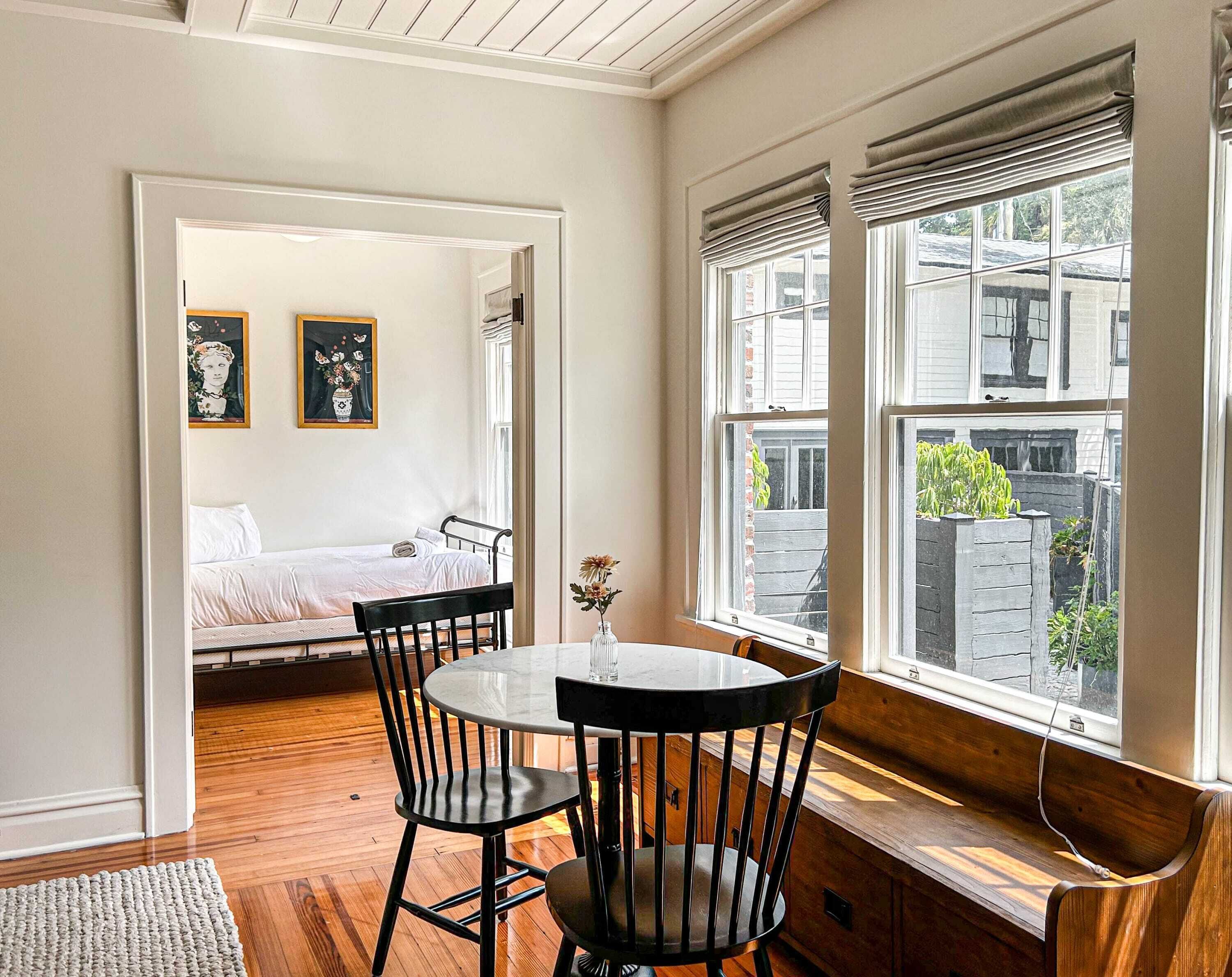 A room with a bed, a dining table with chairs, and a window with a view of the outside.