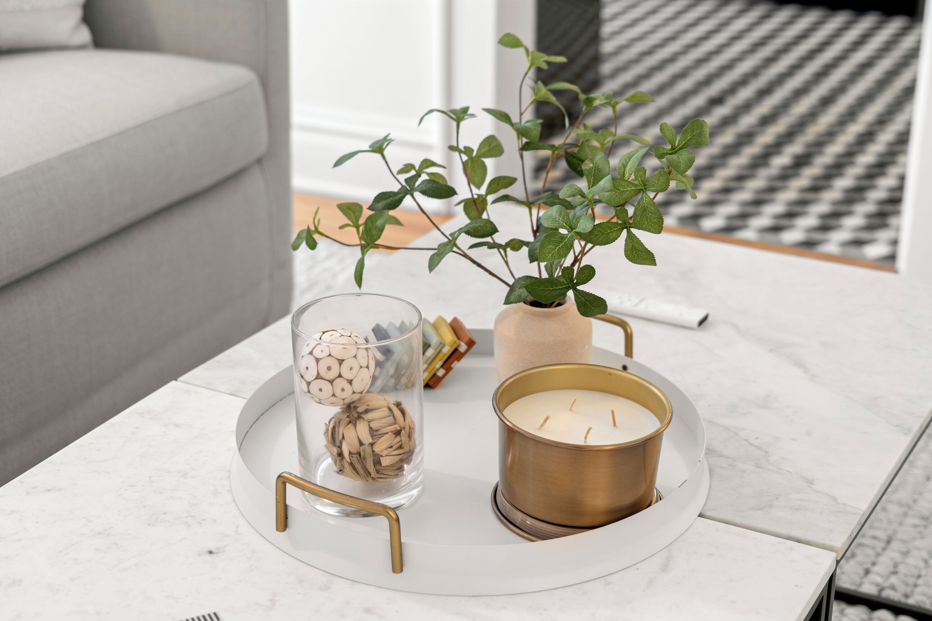 Ein modernes Wohnzimmer mit grauem Sofa, einem Couchtisch aus weißem Marmor, einer Topfpflanze, einer Kerze in einem goldenen Halter und dekorativen Elementen, darunter eine Glasverzierung und eine Holz-Kugel.