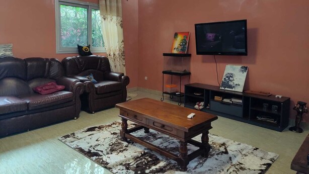 A living room with a leather sofa, a wooden coffee table, a TV on the wall, and a window with curtains.
