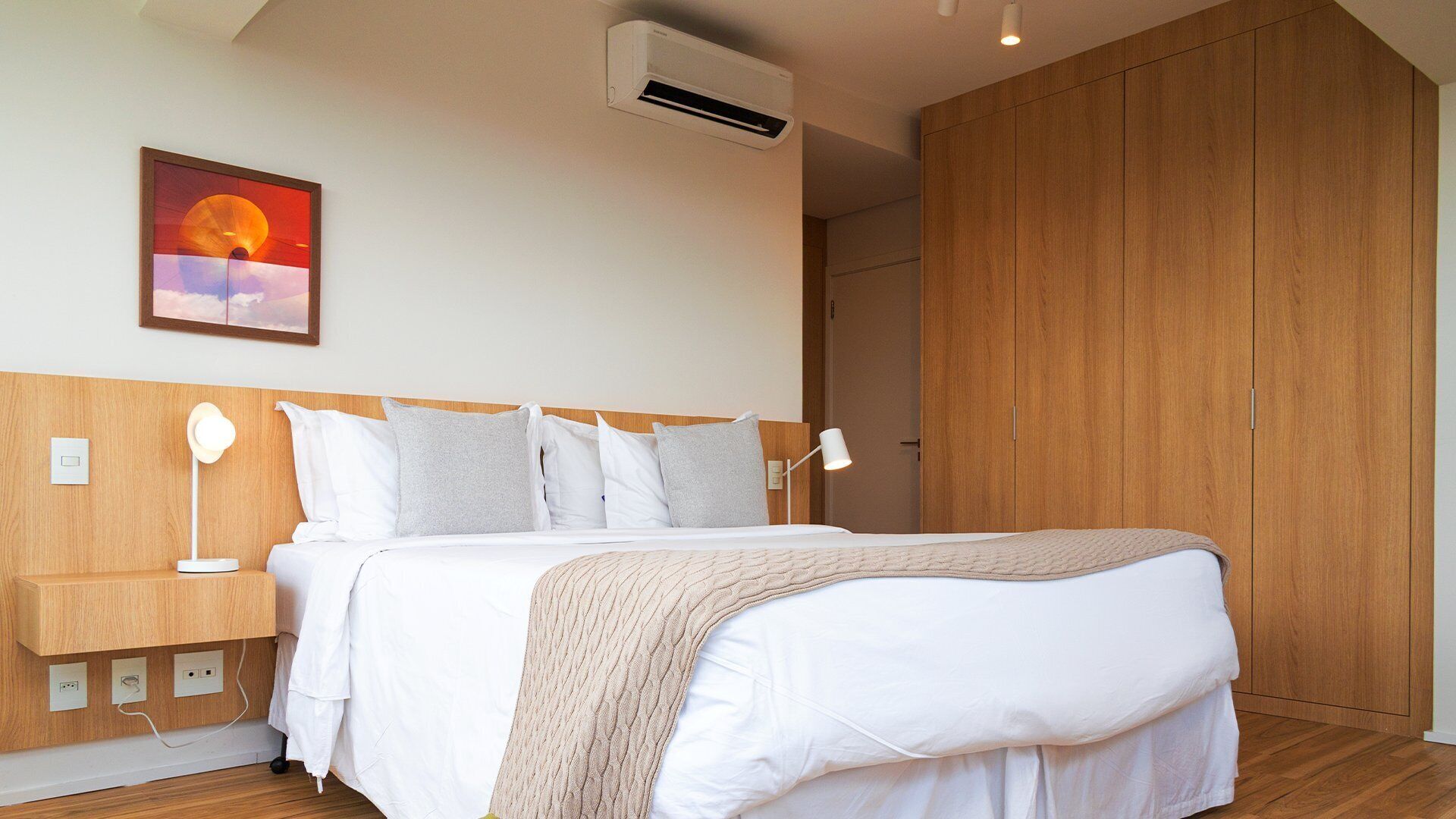 A bedroom with a bed, bedside tables, a lamp, a wall-mounted air conditioner, and a wooden wardrobe.