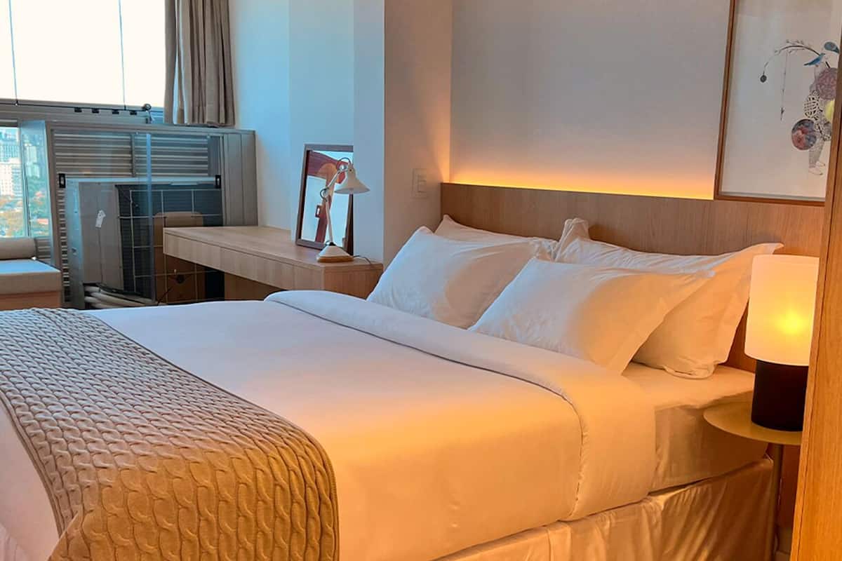 Una camera d'albergo con un letto grande, un comodino con una lampada, una finestra vista sugli edifici e un'opera d'arte appesa al muro.