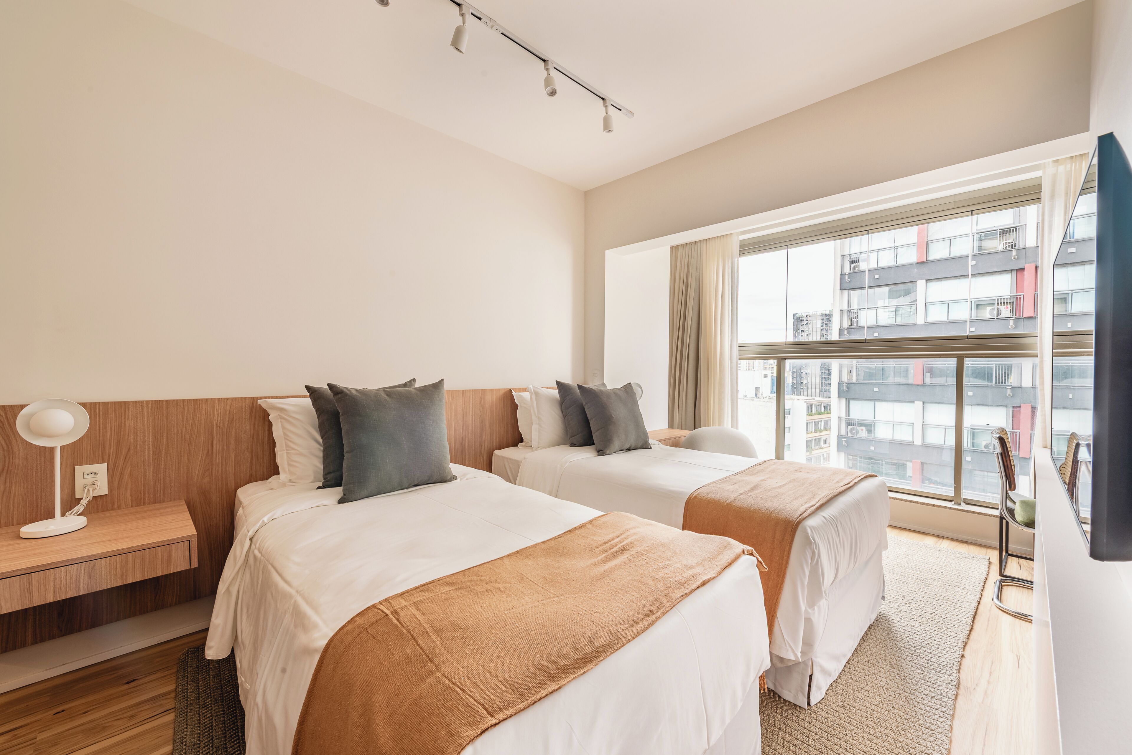 A hotel room with two beds, a desk, a lamp, and a large window with a view of buildings.