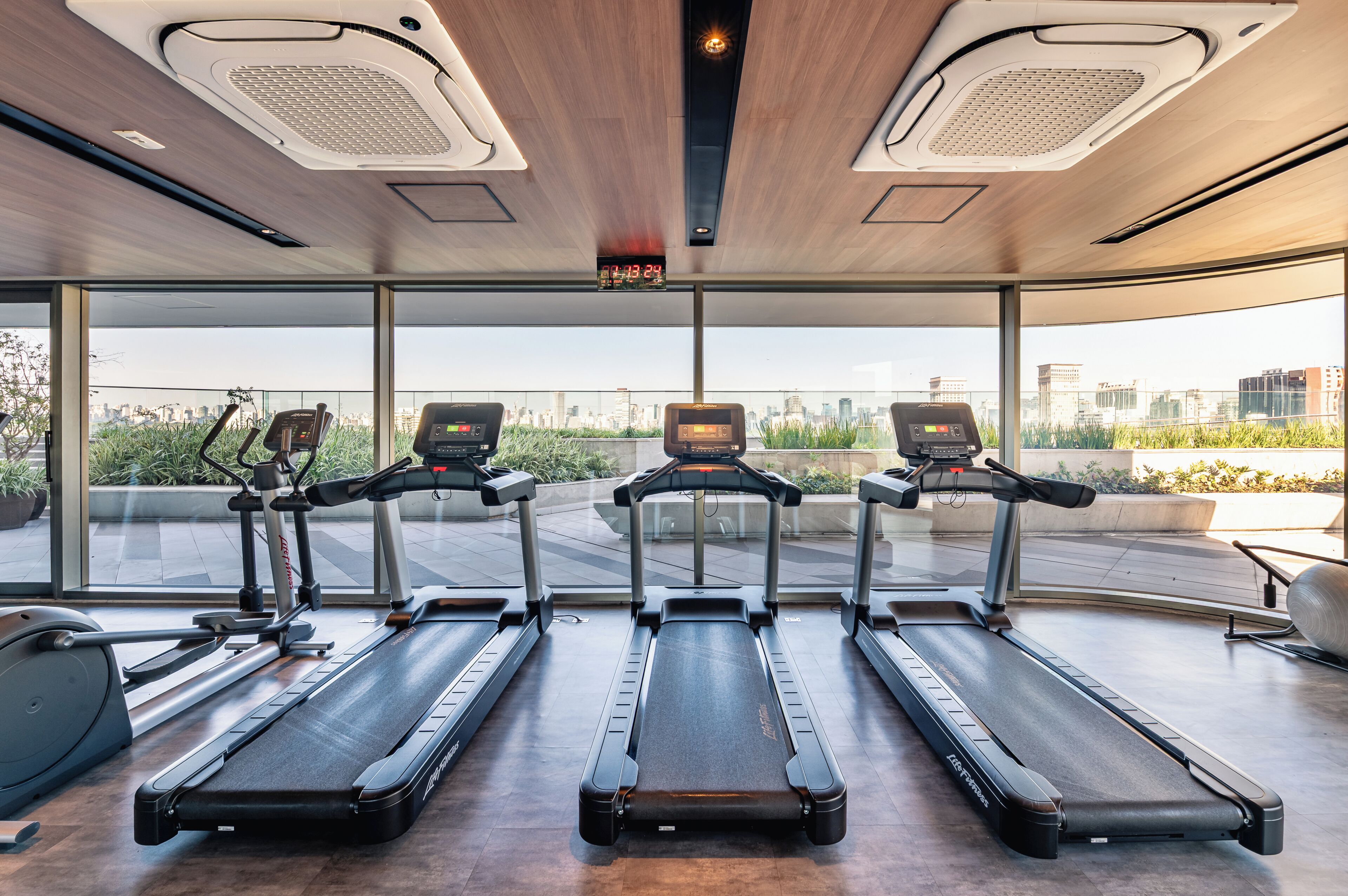 A gym with treadmills, a digital clock, and a city view through large windows.