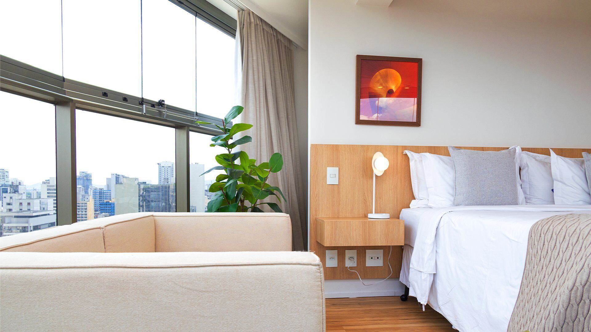 Habitación de hotel con cama, mesita de noche, lámpara y un ventanal con vistas a la ciudad.