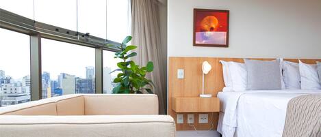 Habitación de hotel con cama, mesita de noche, lámpara y un ventanal con vistas a la ciudad.