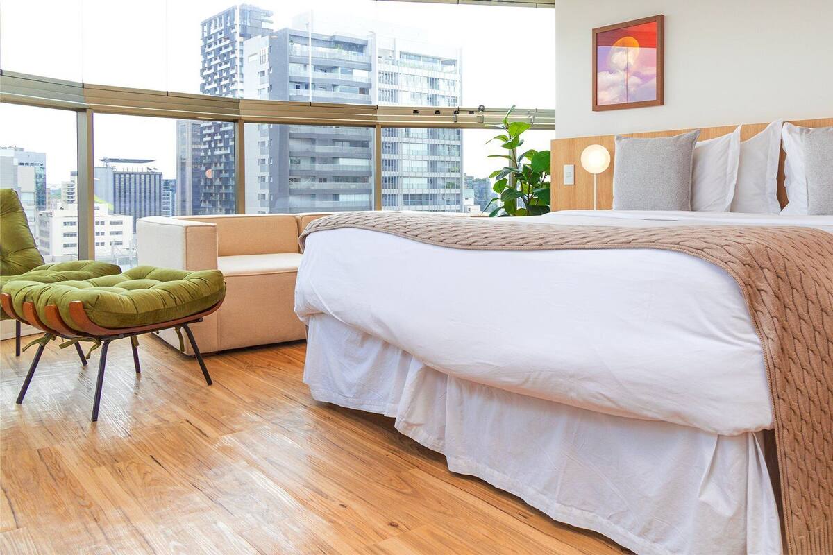 Una camera d'albergo moderna con un letto grande, una poltrona verde e vista sullo skyline urbano, con una finestra che si affaccia su edifici alti.