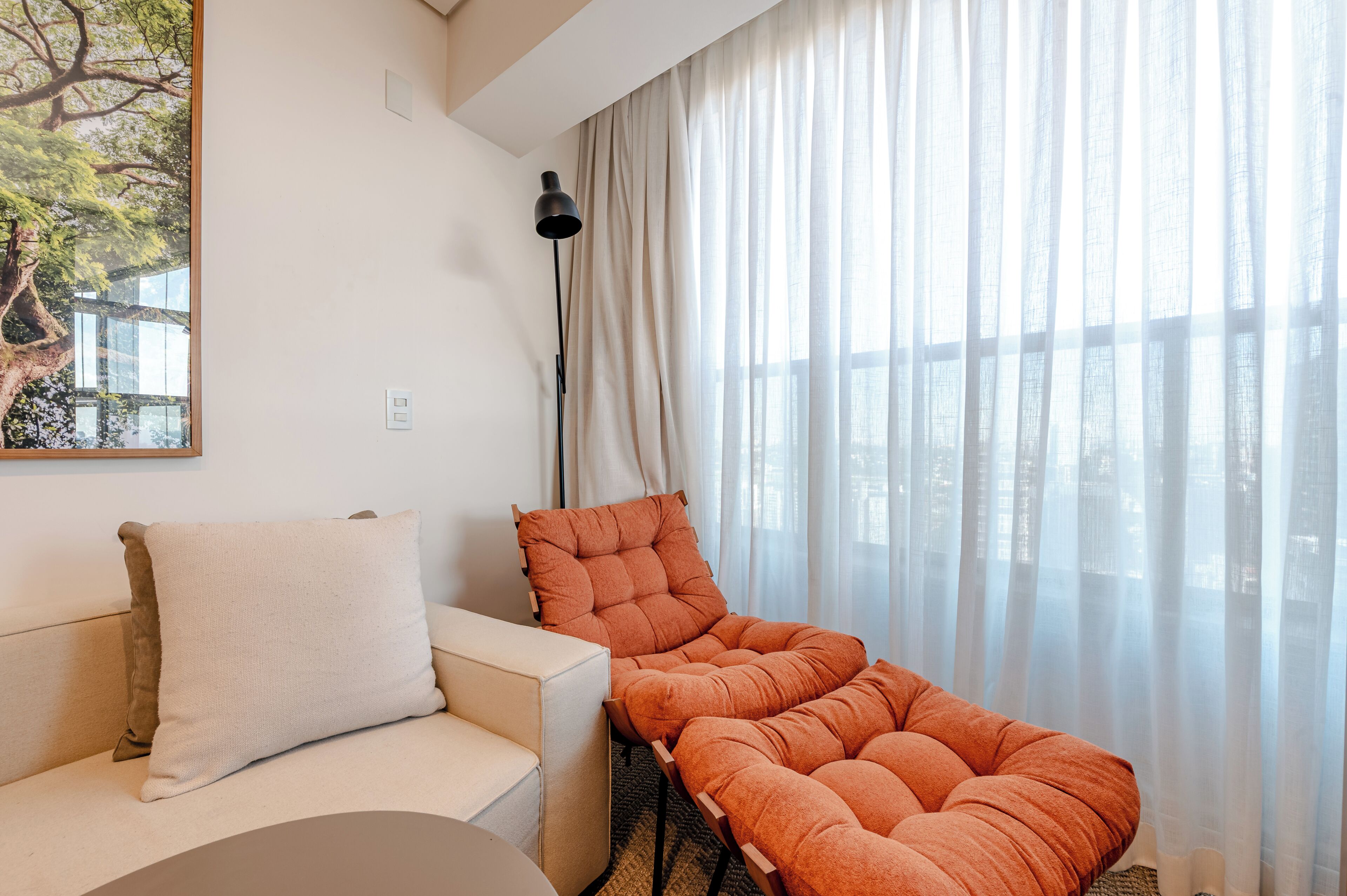 A modern living room with a sofa, a cushioned chair, a floor lamp, and a large window with sheer curtains.