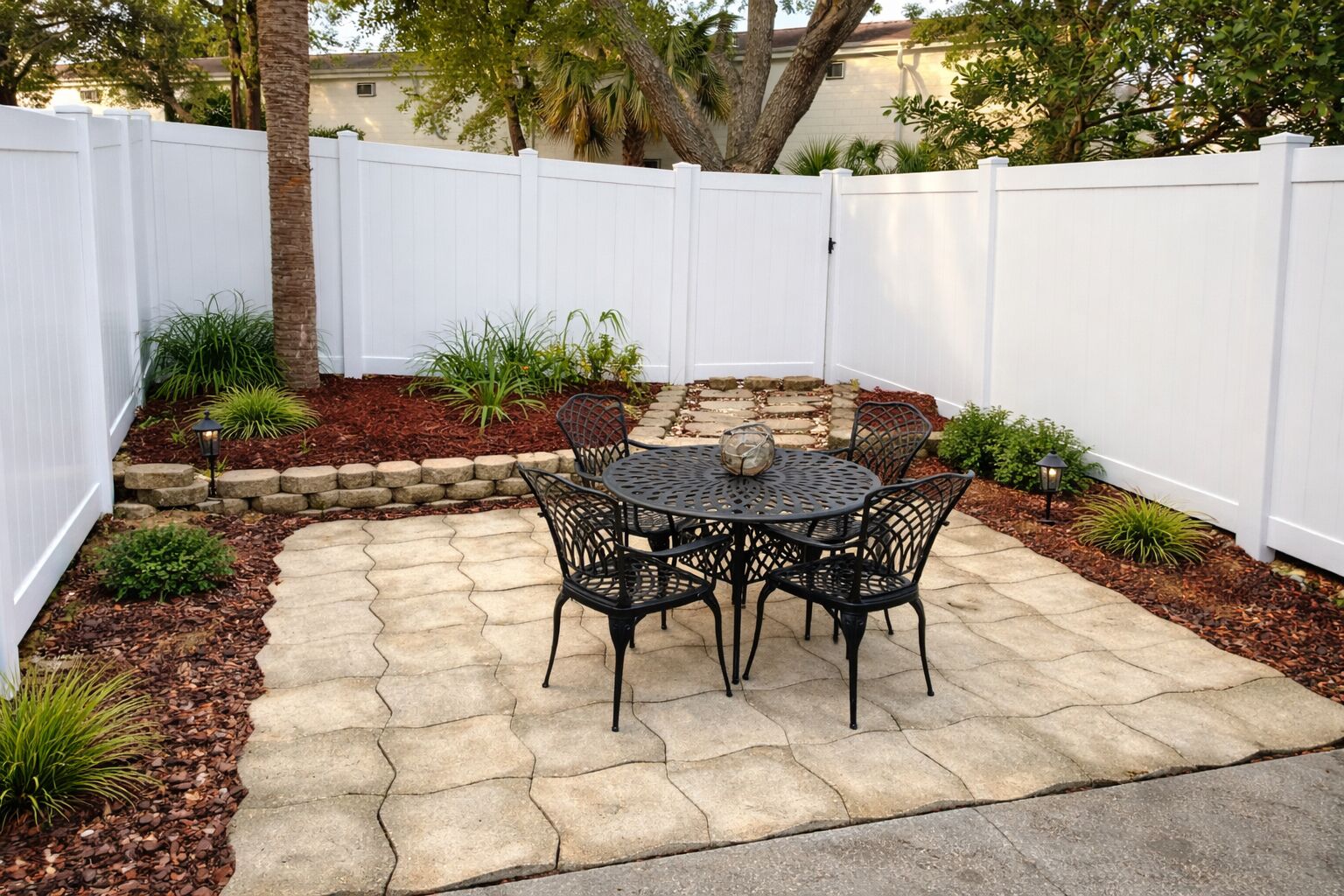 A patio with a round table and chairs, surrounded by a white fence, with plants and mulch around the base.