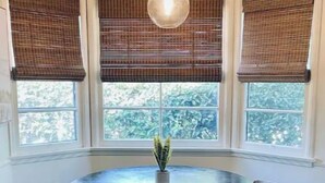 A dining area with a round table, four chairs, a hanging light fixture, and three windows with blinds.